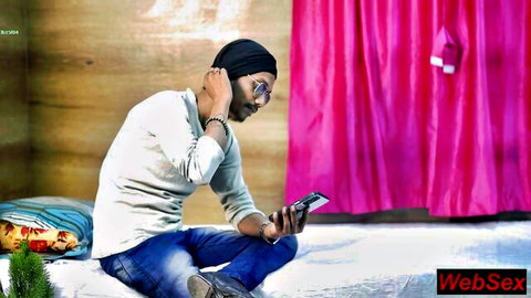 Bearded Indian man in black beanie sitting on bed checking phone with pink curtains