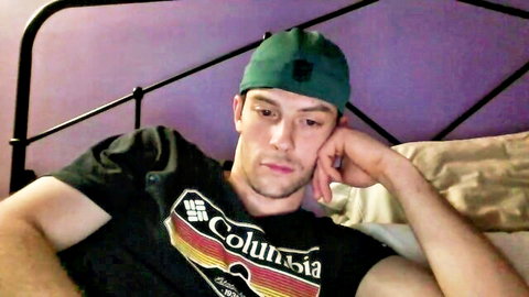 Young white guy in green beanie and Columbia tee lounging thoughtfully on bed, gay amateur thumbnail