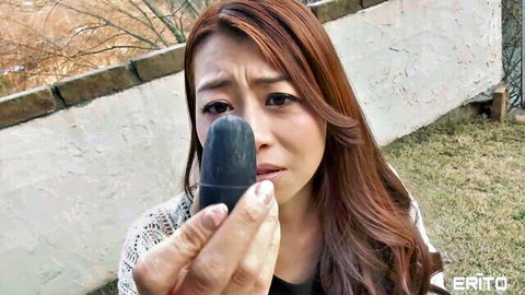 Teary-eyed Japanese babe with long hair holds black dildo to puckered lips outdoors