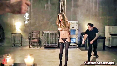 Bound topless submissive babe in black thong stockings dominated by two men in BDSM dungeon