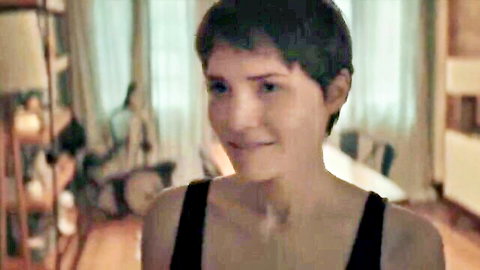 Short-haired brunette smiles seductively in low-cut black tank top showing deep cleavage in bedroom