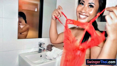 Nude Thai woman with big booty smiling in mirror holding red lingerie