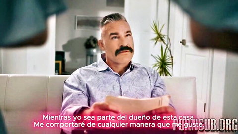 Mature Latino man with mustache sits on couch in patterned shirt speaking Spanish in adult video