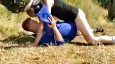 Couple in outdoor grapple on grass, trucker in blue shirt under woman in shorts