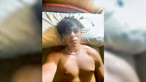 Hot young shirtless Latino hunk lying on bed showing muscular bare chest selfie