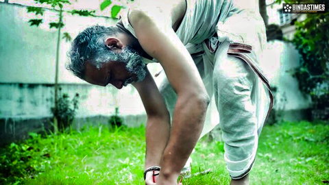 Elderly Indian man with gray beard bending over in white dhoti in lush green garden