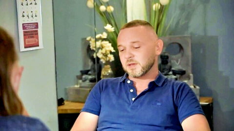 Buzzcut man with goatee relaxes in blue polo shirt as brunette woman faces him in spa room