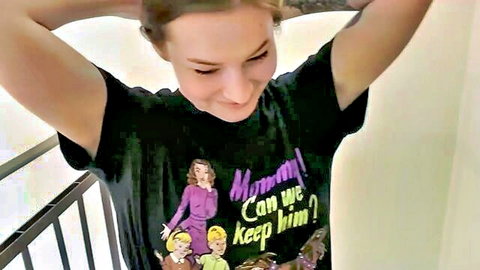 Smiling brunette neighbor in graphic tee, arms raised, teasing public blowjob risk
