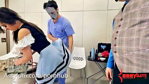 Brunette in off-shoulder top and blue skirt bends over table as masked doctor and suited man watch in exam room