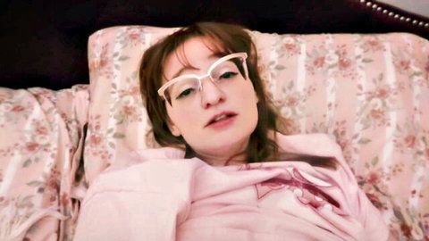 Seductive brunette with glasses lying on bed in pink hoodie, teasing gaze