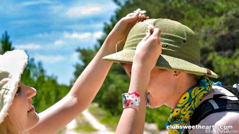 Two smiling teen girls adjusting hats in sunny outdoor forest setting