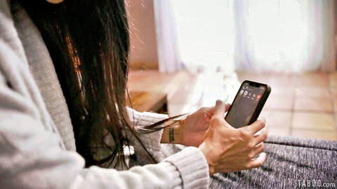 Asian woman with long black hair in gray sweater holding smartphone on couch
