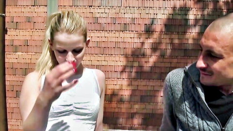 Blonde babe in tight white tank top smoking cigarette with bald guy outdoors against brick wall