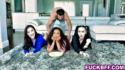 Three sexy teens lying propped on elbows with shirtless muscular guy on fluffy rug holding popcorn