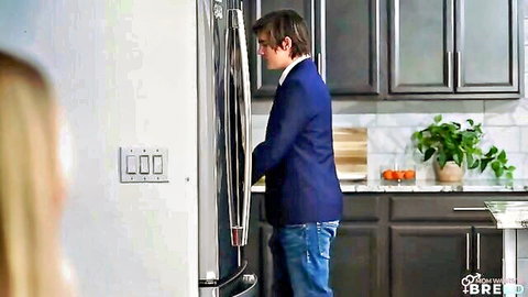 Young man in blue suit opens fridge while blonde stepmom watches in kitchen