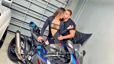 Kelsi Monroe in sheer black bodysuit embracing muscular man by motorcycle