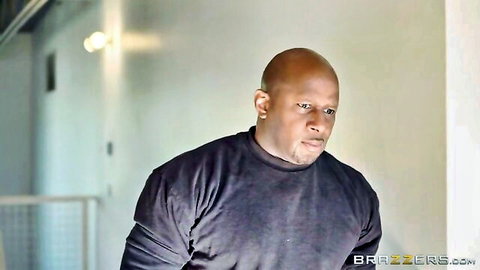 Muscular bald black man in turtleneck, intense gaze in hallway scene