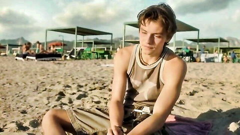 Young man in camo tank top sitting thoughtfully on sunny beach