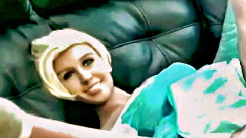 Blonde camgirl in green dress smiling seductively on couch