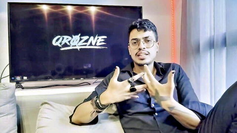 Man in glasses making hand signs on couch, OROZENE TV background
