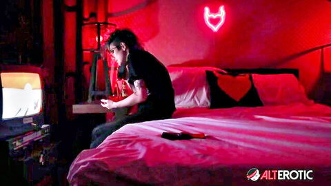 Tattooed emo guy with long hair sits on pink bed playing games in neon red bedroom