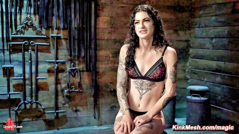 Tattooed woman in black lace lingerie with pained expression in BDSM scene