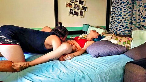 Tattooed woman in black tank top straddles lingerie-clad babe in red bra on bed