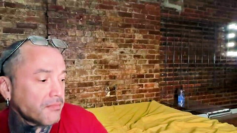 Tattooed man in red shirt with sunglasses on head sits by yellow bed in brick wall room