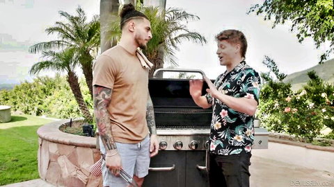 Two tattooed muscular men chatting at outdoor BBQ grill fully clothed