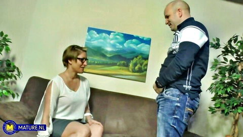 Mature woman with glasses in short skirt sits on couch teasing bald man standing over her in living room