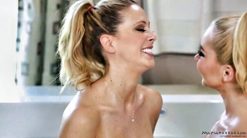 Two nude blonde babes with ponytails smiling intimately in steamy bathtub during lesbian massage