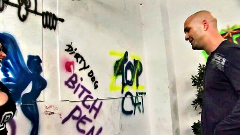 Bald man smiles confidently in front of explicit graffiti wall reading dirty bitch cat