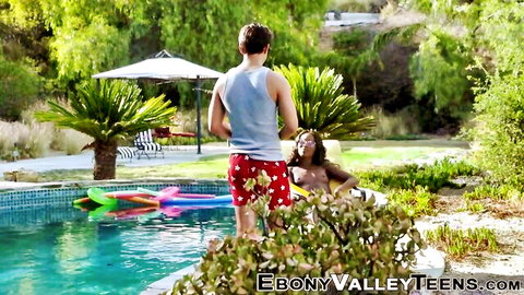 Topless ebony babe with big dark tits smiles up at white stud in red polka-dot trunks by pool