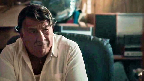 Mature man with chest hair in open white shirt sits intently on leather chair in cozy room
