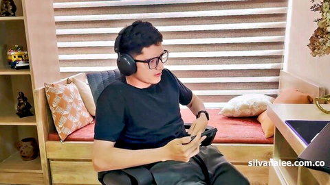 Young Asian guy with glasses and headphones playing video games on controller in cozy bedroom