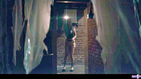 Skinny long-haired brunette in crop top and shorts poses seductively in basement dungeon teasing her medium tits