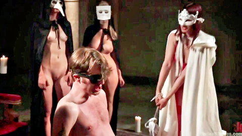 Femdom cult mistress in white cloak torments blindfolded nude slave with masked women