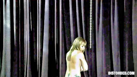 Naked blonde BDSM slave chained by wrists from ceiling showing bare back and tattooed ass