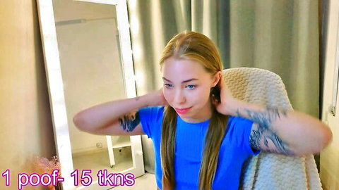 Tattooed blonde teen in blue shirt touches neck smiling on webcam