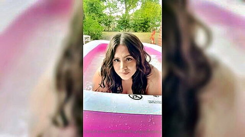 Curvy brunette smiling topless in pink inflatable pool