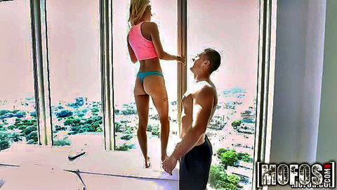 Petite blonde in pink crop top and blue thong stands by window with muscular man behind her
