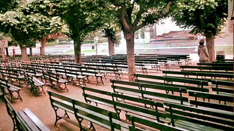 Vintage retro park scene with rows of empty green benches under leafy trees and distant woman walking