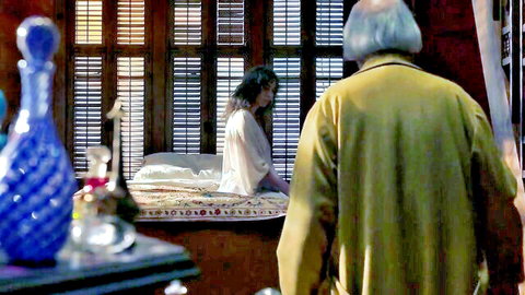 Elderly bald man in yellow jacket faces young brunette in sheer white robe sitting on bed