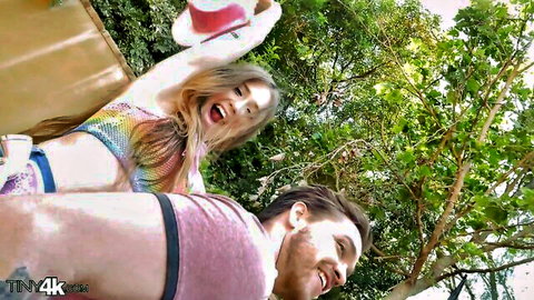 Excited petite blonde in skimpy rainbow bikini top holds hat high as bearded guy grabs her hips from behind outdoors