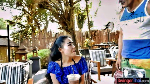 Thai wife in blue off-shoulder top sips drink with man outdoors