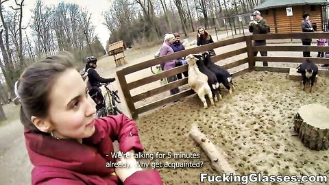 Brunette woman in red jacket and helmet stands by goat enclosure outdoors with people and bikes
