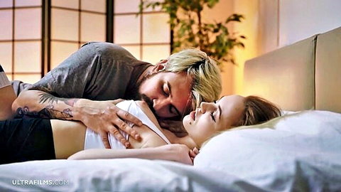 Tattooed blonde guy spoons skinny brunette babe on bed, hand cupping her tits intimately