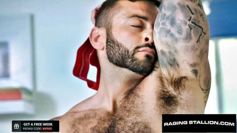 Tattooed muscular man with beard raises arm in red towel, seductive pose