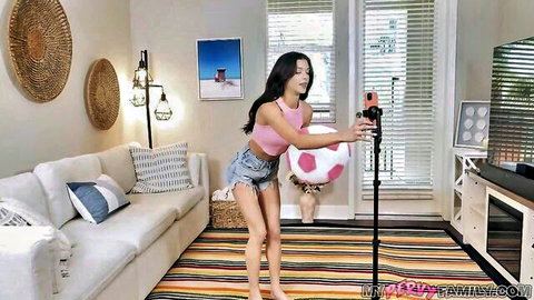 Brunette teen in pink crop top and denim shorts holding soccer ball for playful POV selfie