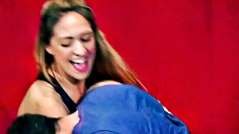 Sexy brunette in black top smiles wide while embraced from behind by man in blue shirt on red stage.